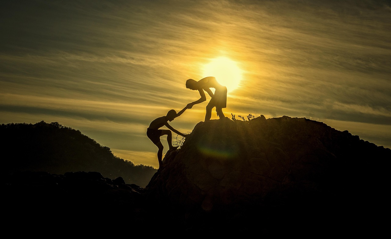 One person reaches down to help another climb up a hill at sunset