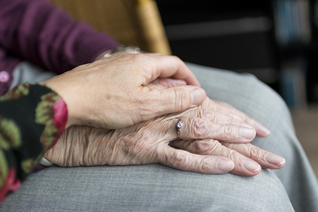 A younger hand rests gently on the clasped hands of an elderly person, offering comfort